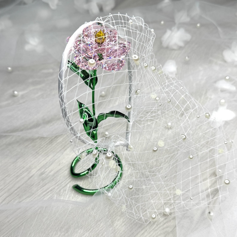 May include: A white headband featuring a pink crystal flower with a yellow centre, green metal stem and leaves, and a white netting veil adorned with small white pearls. The base of the stem forms a circular green metal loop for stability.