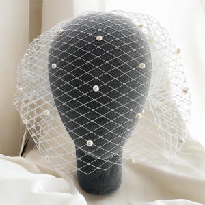 May include: A white birdcage veil featuring a diamond-shaped netting pattern adorned with evenly spaced, small round white pearls throughout the mesh. The veil is draped over a dark grey mannequin head, highlighting the contrast between the white netting and the dark form underneath.