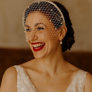 May include: A smiling person wearing a white birdcage veil adorned with pearls. The person is wearing a white dress and gold hoop earrings with a pearl. The person has red lipstick.
