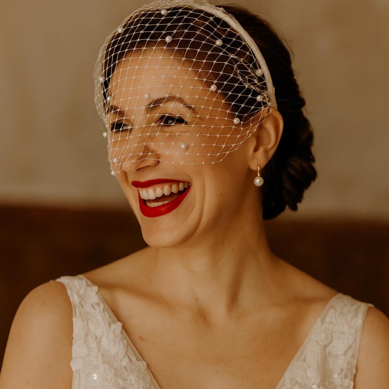 May include: A white birdcage veil headband featuring a delicate netting pattern adorned with small, evenly spaced white pearls. The accessory is designed to sit across the upper face, providing a structured yet lightweight appearance for formal headwear.