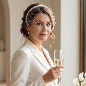 May include: A white birdcage veil headband featuring a delicate netting pattern adorned with small, evenly spaced white pearls. The veil is attached to a smooth, solid white headband, designed to sit across the upper forehead and eyes.