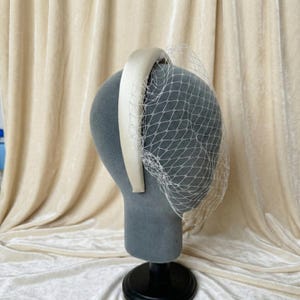Op de afbeelding: Een cr&egrave;mekleurige haarband met bijpassende sluier, tentoongesteld op een grijze mannequinhoofd. De haarband is breed en satijnachtig, en de sluier is een fijn, netachtig materiaal. De achtergrond is een zachte, neutrale stof.