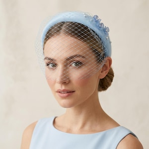 Könnte beinhalten: Ein hellblaues, gepolstertes Stirnband mit einem zarten Birdcage-Schleier, der das obere Gesicht bedeckt. Das Stirnband ist mit kleinen hellblauen Blumenakzenten und winzigen weißen Perlen verziert, die seitlich auf der glatten Stoffoberfläche sitzen.
