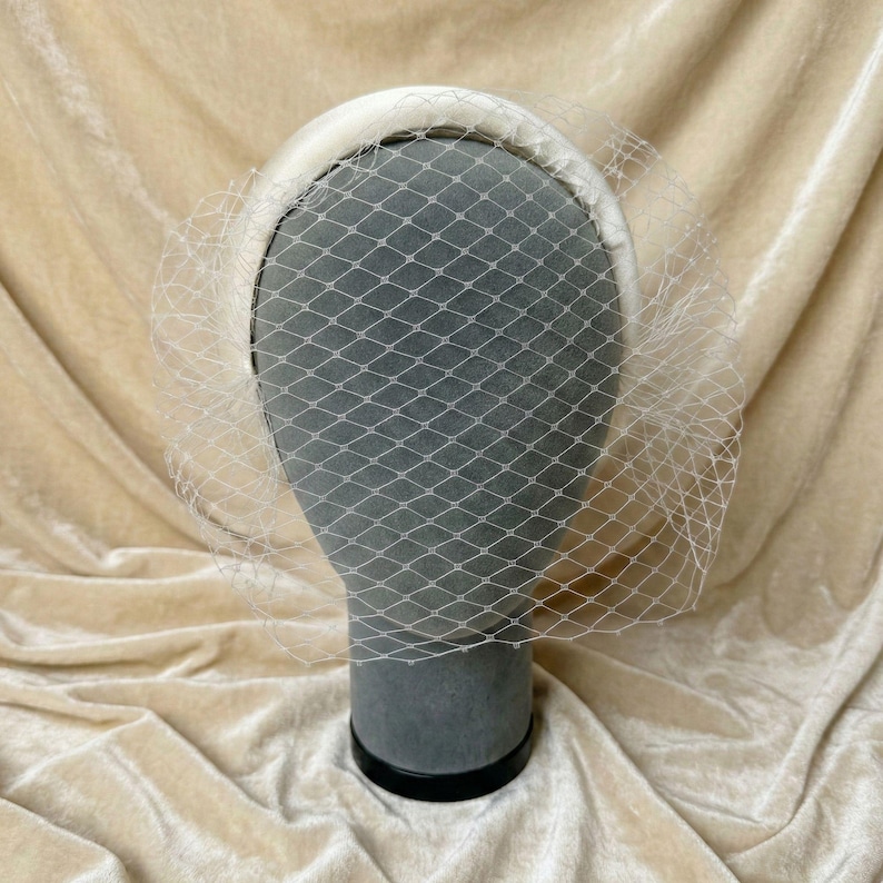 Op de afbeelding: Een cr&egrave;mekleurige haarband met bijpassende sluier. De sluier is een fijn, wit net dat over de voorkant valt. De haarband staat op een grijze mannequinhoofd, tegen een beige stoffen achtergrond.