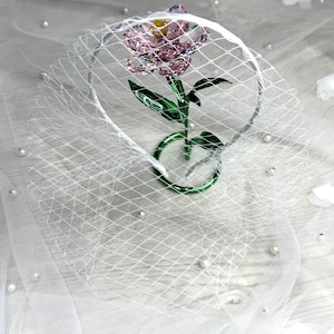 May include: A headband featuring a white birdcage veil and a faceted pink crystal flower with a yellow centre and green crystal stem and leaves. The flower is positioned upright within the circular frame of the headband, creating a decorative accessory.