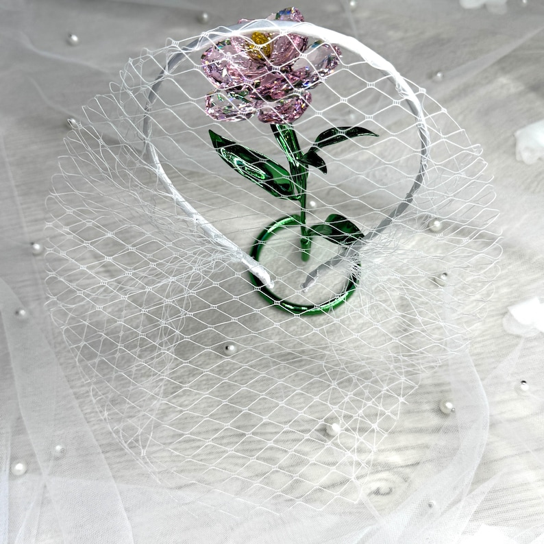 May include: A headband featuring a white birdcage veil and a faceted pink crystal flower with a yellow centre and green crystal stem and leaves. The flower is positioned upright within the circular frame of the headband, creating a decorative accessory.