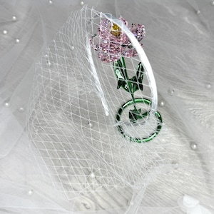 May include: A white headband featuring a white birdcage veil and a decorative pink flower ornament with a yellow centre, attached to a green stem and circular base. The flower petals are faceted and translucent, reflecting light with a sparkling effect.