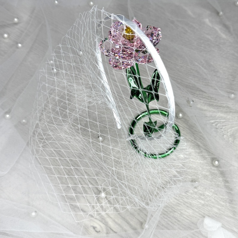 May include: A white headband featuring a white birdcage veil and a decorative pink flower ornament with a yellow centre, attached to a green stem and circular base. The flower petals are faceted and translucent, reflecting light with a sparkling effect.