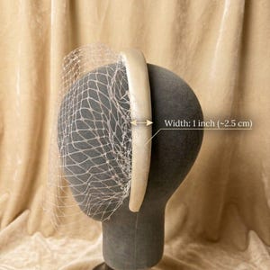 Op de afbeelding: Een cr&egrave;mekleurige haarband met een delicate netsluier. De haarband is 2,5 cm breed. De sluier is aan de haarband bevestigd en valt naar beneden. Het hoofddeksel wordt op een mannequinhoofd getoond.