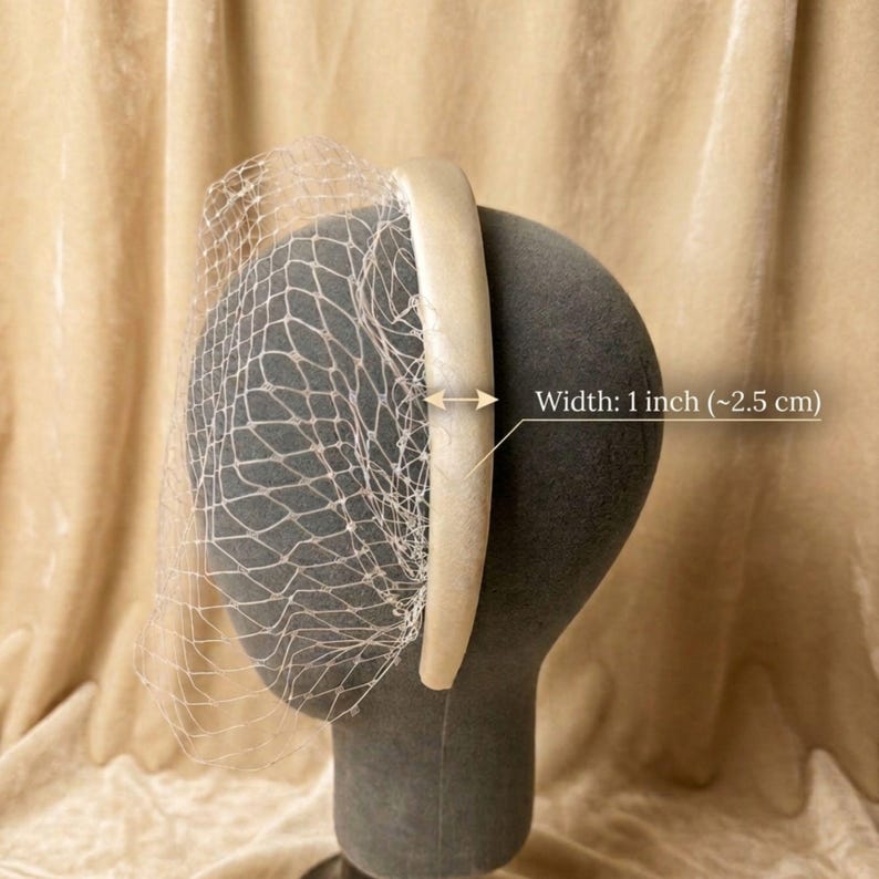Op de afbeelding: Een cr&egrave;mekleurige haarband met een delicate netsluier. De haarband is 2,5 cm breed. De sluier is aan de haarband bevestigd en valt naar beneden. Het hoofddeksel wordt op een mannequinhoofd getoond.
