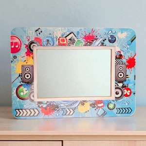 Puede incluir: Un espejo rectangular con un marco azul claro decorado con gráficos coloridos con temas musicales, que incluyen notas musicales, altavoces y salpicaduras de pintura. El espejo está sobre una superficie de madera clara contra una pared azul pálido.