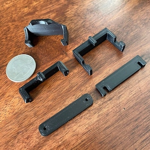 May include: An assortment of black 3D-printed plastic components, including several L-shaped brackets, a rectangular piece, and a curved piece. A US quarter is included for size reference. The items are arranged on a wooden surface.