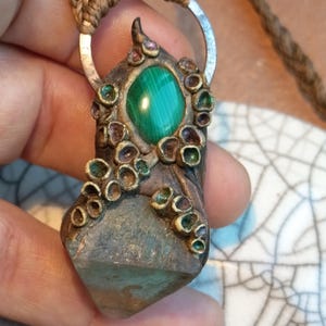 May include: A handcrafted pendant necklace featuring a green malachite stone set in a decorative metal casing. The pendant has a unique design with small, colorful embellishments and is suspended from a braided cord.