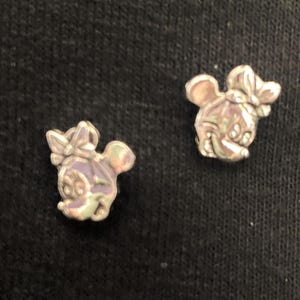 Puede incluir: Dos pendientes de plata con un dije de rat&oacute;n de dibujos animados con un lazo. El rat&oacute;n est&aacute; mirando hacia la derecha en ambos pendientes.