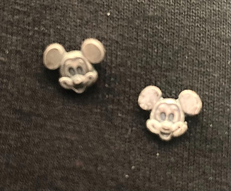 Puede incluir: Dos peque&ntilde;as figuras de pl&aacute;stico de Mickey Mouse de color blanco. Las figuras est&aacute;n mirando hacia adelante con las orejas apuntando hacia los lados. Las figuras est&aacute;n sobre una superficie gris oscuro.