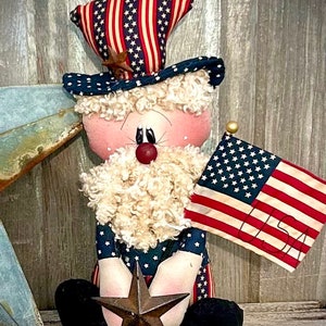May include: A patriotic rag doll dressed in red, white, and blue with a white beard, holding a star and an American flag.