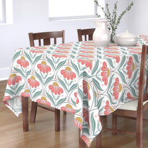 Puede incluir: Un mantel blanco con un estampado floral repetido. El patrón presenta flores de color coral con centros amarillos, hojas verdes y pequeños puntos rosas. El mantel está sobre una mesa de madera.
