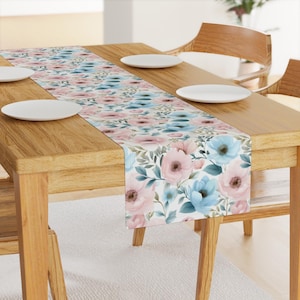 May include: A white table runner with a floral pattern featuring pink and blue flowers on a white background. The runner is placed on a wooden table with two white plates.