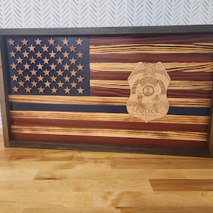 May include: A wooden American flag with a blue line and a police corporal badge in the center. The flag is framed in dark wood.