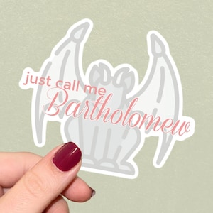 May include: A die-cut sticker featuring a light grey gargoyle illustration with large wings. Overlaid on the gargoyle is the pink text "just call me Bartholomew" written in a combination of a simple sans-serif font and a cursive script font.