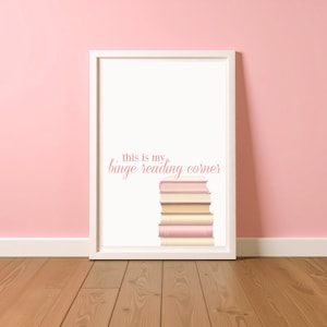 May include: A framed print with the text "this is my binge reading corner" in a pink script font. A stack of books in shades of pink and cream is below the text. The frame is white, and the background is a light pink.