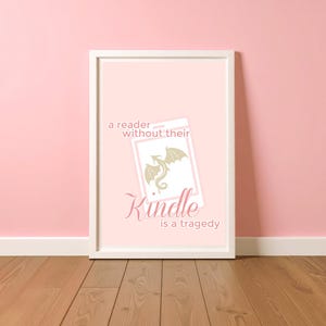 Op de afbeelding: Een ingelijste print met een roze achtergrond en een witte rand. De print toont een gestileerde afbeelding van een Kindle e-reader met een draakafbeelding. De tekst op de print luidt: "een lezer zonder hun Kindle is een tragedie."
