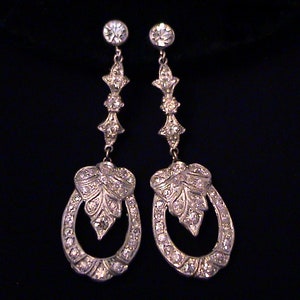 May include: A pair of silver-toned drop earrings featuring a vertical design with a circular stud at the top, a central decorative link, and a large oval hoop at the bottom containing a leaf-shaped element, all encrusted with clear rhinestones.