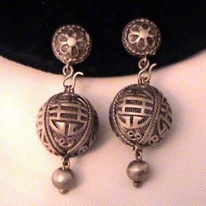 May include: A pair of silver-toned drop earrings featuring a circular stud with a floral pattern, connected by a hook to a spherical charm with an openwork filigree design and a Chinese character, finished with a small dangling silver-toned bead.