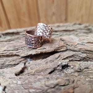May include: Two wide copper rings featuring a hammered, dimpled texture across the entire outer surface. The metal has a warm, reddish-brown metallic finish with dark oxidation in the recessed areas of the indentations, creating a high-contrast look.