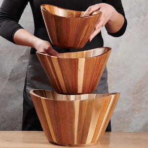 May include: Set of three wooden salad bowls. The bowls are made of light and dark brown wood, with a curved, boat-like shape. The bowls are stacked on top of each other, with a person holding the top bowl.