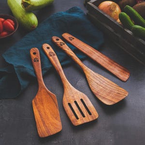 May include: Four wooden kitchen utensils, including a spatula and slotted spoon, are arranged on a dark surface. The utensils are made of a warm-toned wood and feature a decorative design on the handles. Fresh vegetables are in the background.