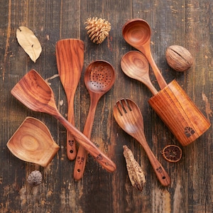 May include: A collection of wooden kitchen utensils, including a spatula, slotted spoon, ladle, and serving fork, arranged on a dark wood surface. The utensils are a warm brown color and feature a decorative design on the handles. A wooden utensil holder is also present.