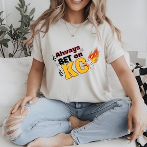 May include: A white t-shirt with a graphic design that says "Always Bet on KC" with a yellow and black colour scheme and a red and yellow flame design.