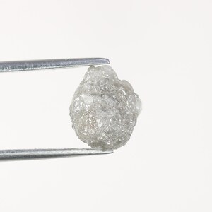 Op de afbeelding: Een ruwe, ongeslepen, lichtgrijze diamant die door een zilveren pincet wordt vastgehouden. De diamant heeft een getextureerd, onregelmatig oppervlak en een troebel uiterlijk. De achtergrond is effen wit.