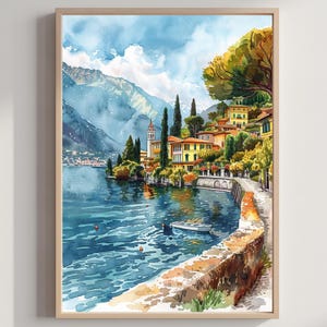 May include: Watercolour painting of a lakeside village with colourful buildings, trees, and mountains in the background. A stone wall lines the water's edge, and a small boat floats on the blue water. The artwork is framed in a light wood frame.