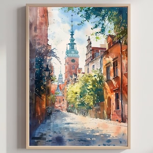 May include: Watercolor painting of a European city street scene. The artwork features buildings in shades of orange and red, a tall clock tower, and a cobblestone street. The sky is blue with clouds, and trees line the street. The artwork is framed in a light wood frame.