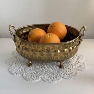 May include: A round brass bowl with two side handles and three small feet, featuring an embossed decorative pattern along the exterior rim. The bowl contains three whole oranges. The metal has a polished, reflective gold-toned finish.
