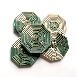 以下が含まれることがあります： 迷路のデザインが施された、5つの八角形のコースターのコレクション。コースターは緑と金色の配色で、中央に昆虫のようなエンブレムがあります。白い背景に配置されています。