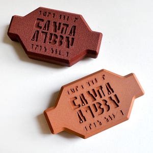 以下が含まれることがあります： 彫刻されたテキストが施された、2つの長方形の赤茶色のプラーク。プラークは、テクスチャ加工された表面と、延長された端を持つユニークな形状をしています。テキストは数字と記号の組み合わせのようです。