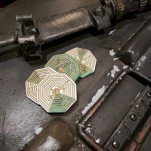 May include: Three octagonal coasters featuring a white and gold labyrinth pattern with a teal green accent section. Each coaster displays a central gold emblem within the maze design, showcasing a layered construction with distinct geometric lines.