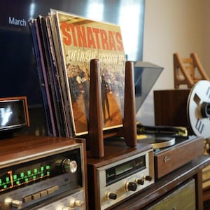 May include: A wooden record holder displaying a vinyl album titled "Sinatra's Swinging Session!!!". The album is propped up on a vintage stereo system with a silver face and wooden accents. Other vinyl records are stacked behind the album.