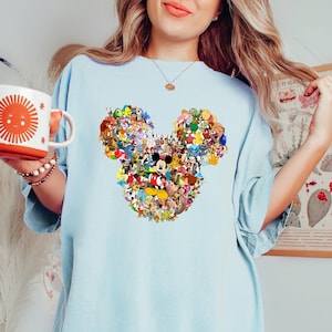 May include: Light blue t-shirt featuring a Mickey Mouse head design composed of various Disney characters. The shirt is a soft, comfortable material. A white mug with a smiling sun design is held in the left hand.