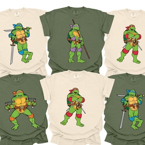 May include: Six t-shirts featuring Teenage Mutant Ninja Turtles characters in various poses. The shirts are in cream and olive green, with each turtle wearing a different colored mask and wielding a weapon. The cartoon characters are in the center of the shirts.