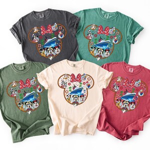 May include: Five colorful t-shirts featuring a Minnie Mouse design. The design includes a cruise ship, various cartoon characters, and a red bow. The shirts are in shades of gray, green, cream, and red.