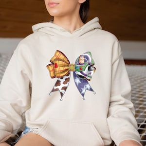 May include: A beige hoodie featuring a colorful bow design. The bow is composed of a plaid section, a cow print section, and a section with a purple star pattern. The bow also features a graphic of Buzz Lightyear.