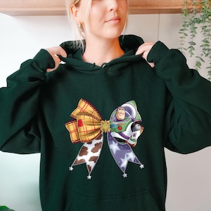 May include: Dark green hoodie featuring a colorful bow design with a Buzz Lightyear graphic. The bow has a plaid pattern, cow print, and a purple ribbon with white stars. The hoodie has a front pocket and a drawstring hood.