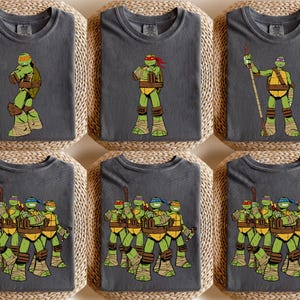 May include: Six charcoal gray t-shirts featuring the Teenage Mutant Ninja Turtles. Each shirt displays a different graphic of the turtles in action poses. The turtles are depicted in their signature colors: green, yellow, brown, and red, with detailed illustrations.