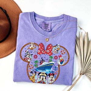 May include: A lilac Comfort Colors crew neck t-shirt with a Minnie Mouse-shaped design featuring Disney characters and a cruise ship. The design includes Mickey Mouse, Minnie Mouse, Donald Duck, and Goofy. A gold necklace and a brown hat are also visible.