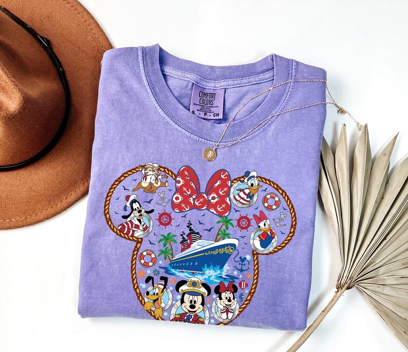 May include: A lilac Comfort Colors crew neck t-shirt with a Minnie Mouse-shaped design featuring Disney characters and a cruise ship. The design includes Mickey Mouse, Minnie Mouse, Donald Duck, and Goofy. A gold necklace and a brown hat are also visible.