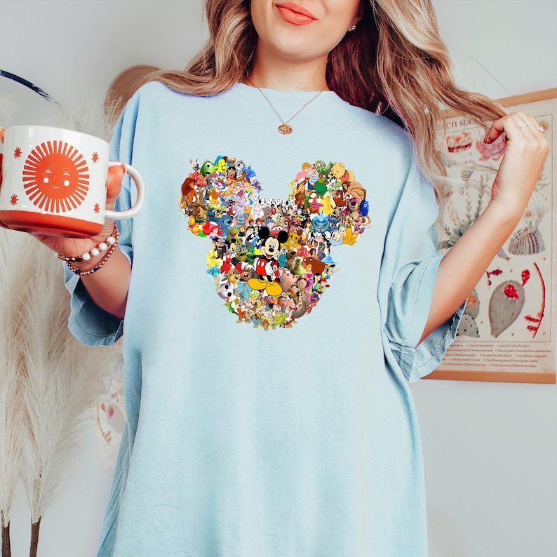 May include: Light blue t-shirt featuring a Mickey Mouse head design composed of various Disney characters. The shirt is a soft, comfortable material. A white mug with a smiling sun design is held in the left hand.